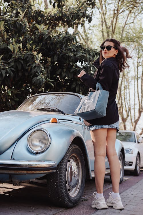 A woman in shorts and a jacket is standing next to a vintage beetle
