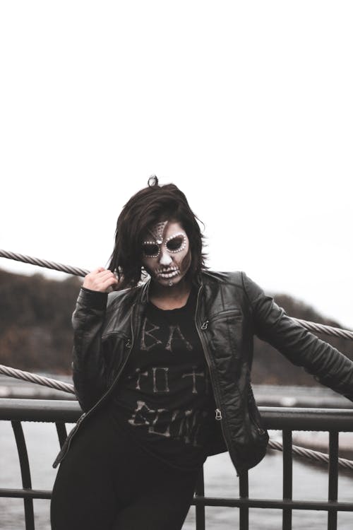 бесплатная Женщина в серой маске Хэллоуина и черной кожаной куртке стоит у черных перил Стоковое фото