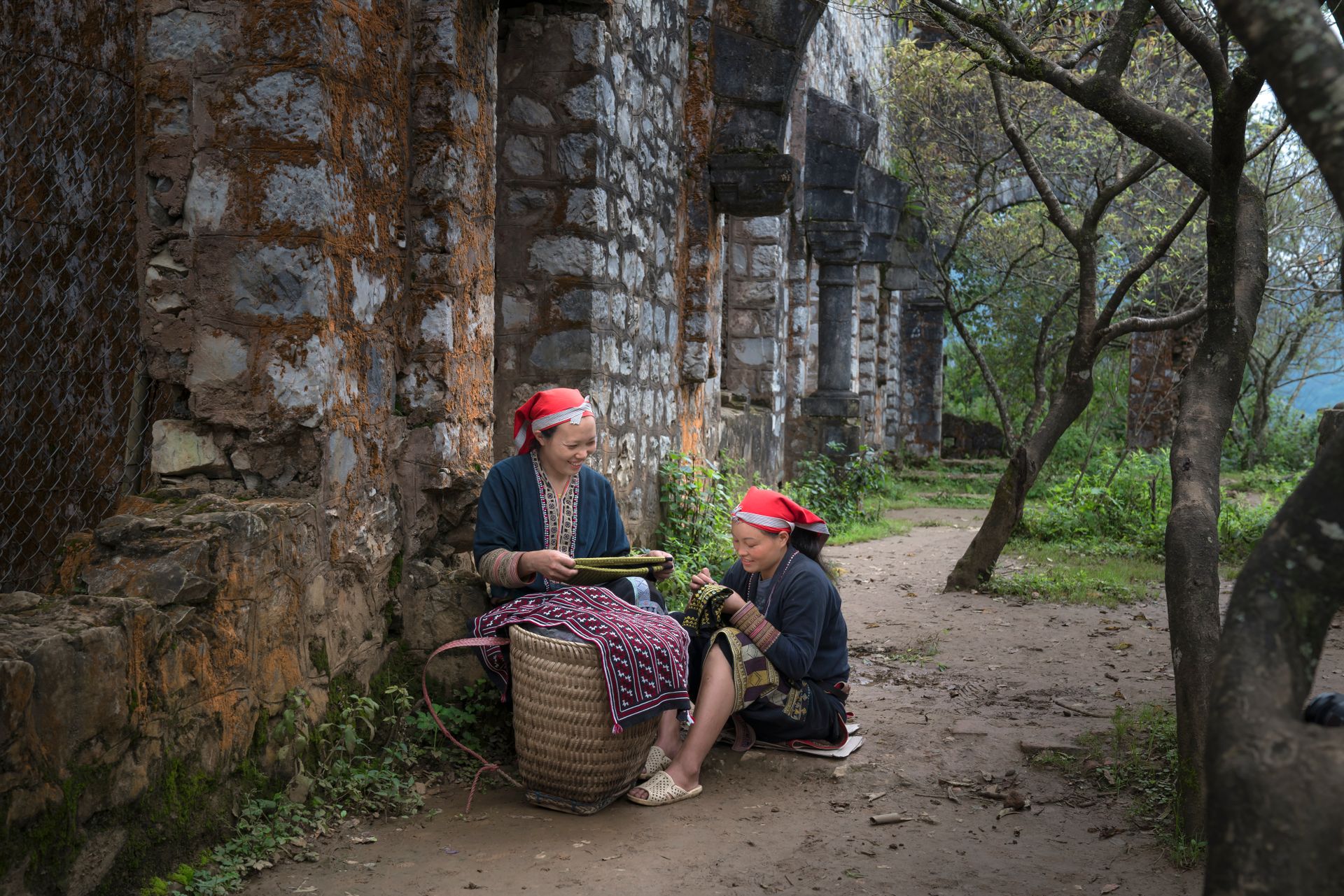 Two women in traditional attire weaving baskets outdoors near a stone building, showcasing cultural craftsmanship.