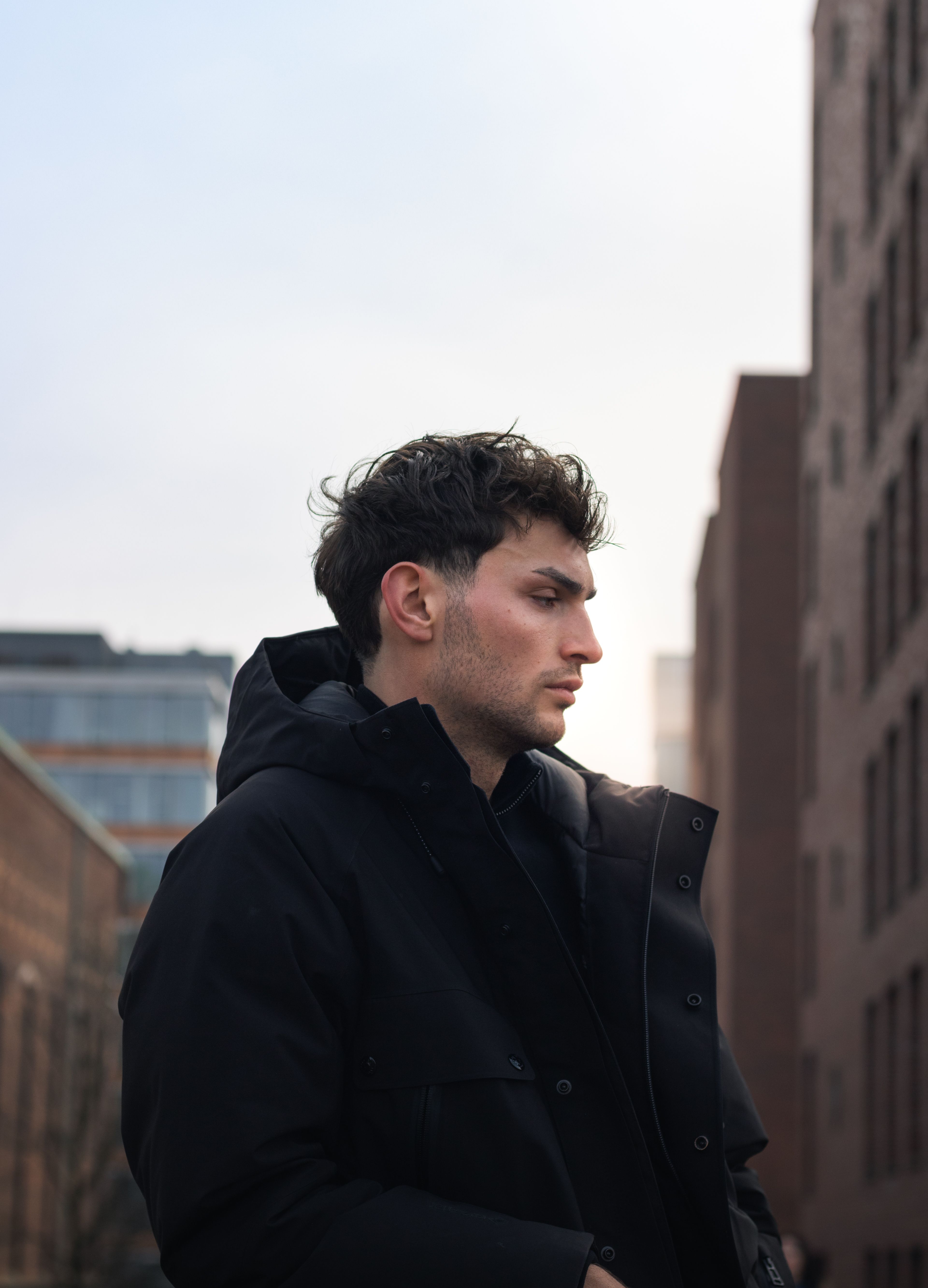 Young man in black jacket posing in urban setting, showcasing modern fashion style.