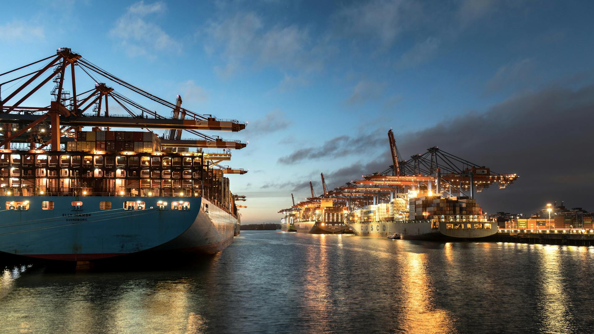 Cargo ships docked at Hamburg harbor, illuminated under evening sky. A hub of logistics and commerce.