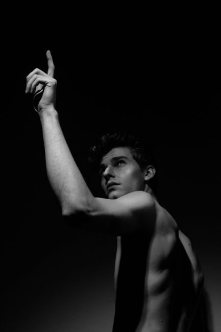 Black and white portrait of a shirtless man pointing up, captured in dramatic lighting.