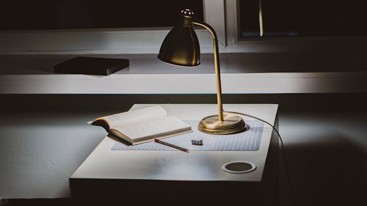 Powered-on Gold Desk Lamp On Desk With Opened Book