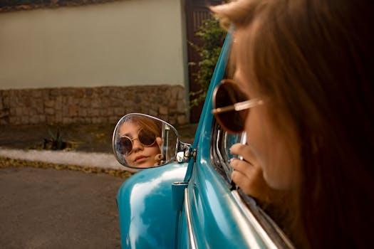 A woman wearing sunglasses reflects in a classic car's side mirror, creating a stylish portrait.