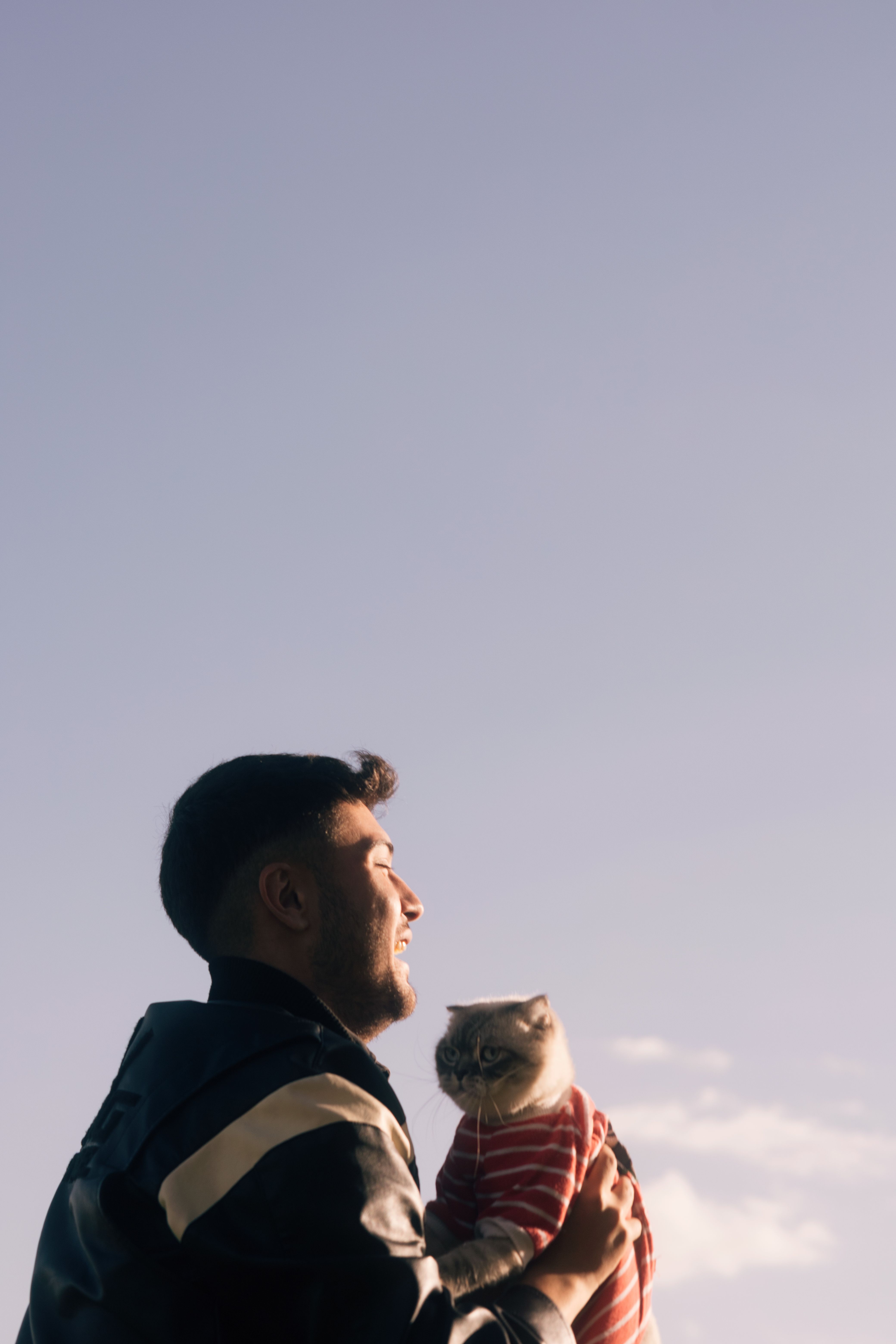 A man smiling while holding a cat in a striped sweater, clear sky background.