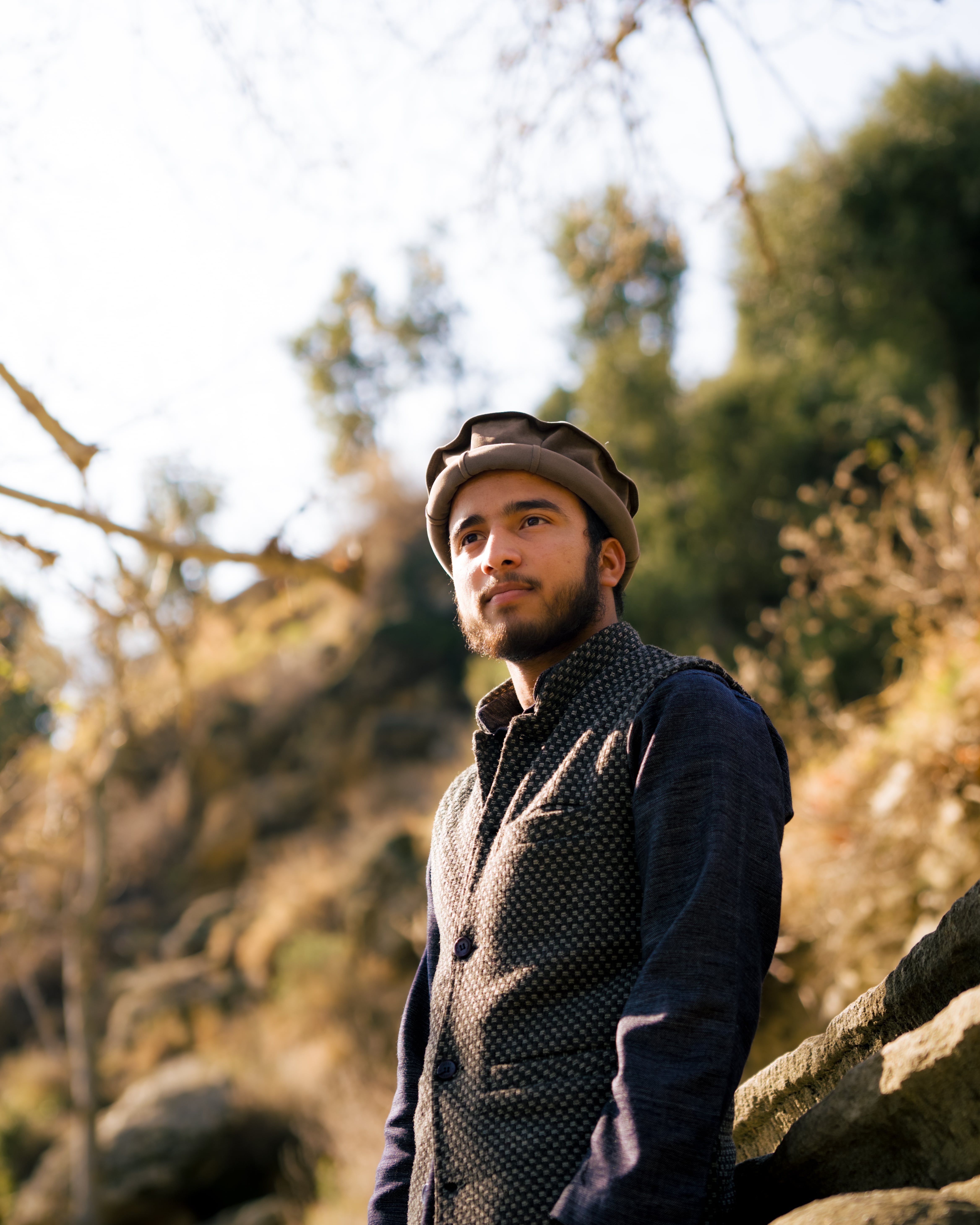 A man with a beard and hat stands outdoors in a natural, rural setting, showcasing fashion.