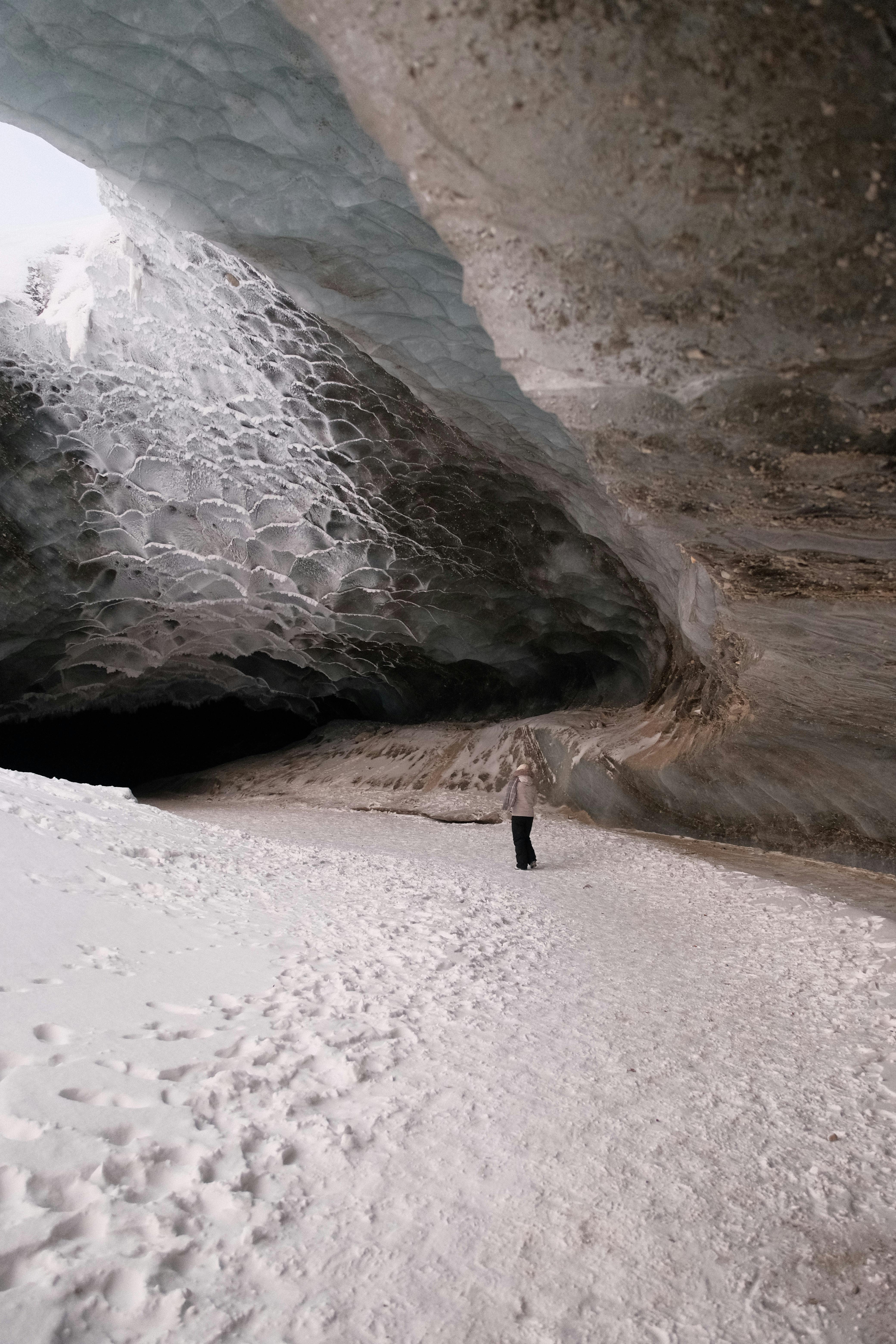 A lone figure stands inside a vast, stunning ice cave with textured icy walls.