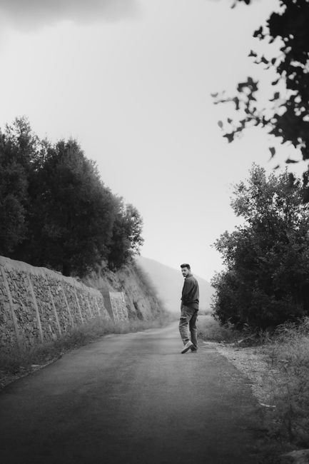 A lone man walks on a rural path lined with trees and shrubs under a cloudy sky.