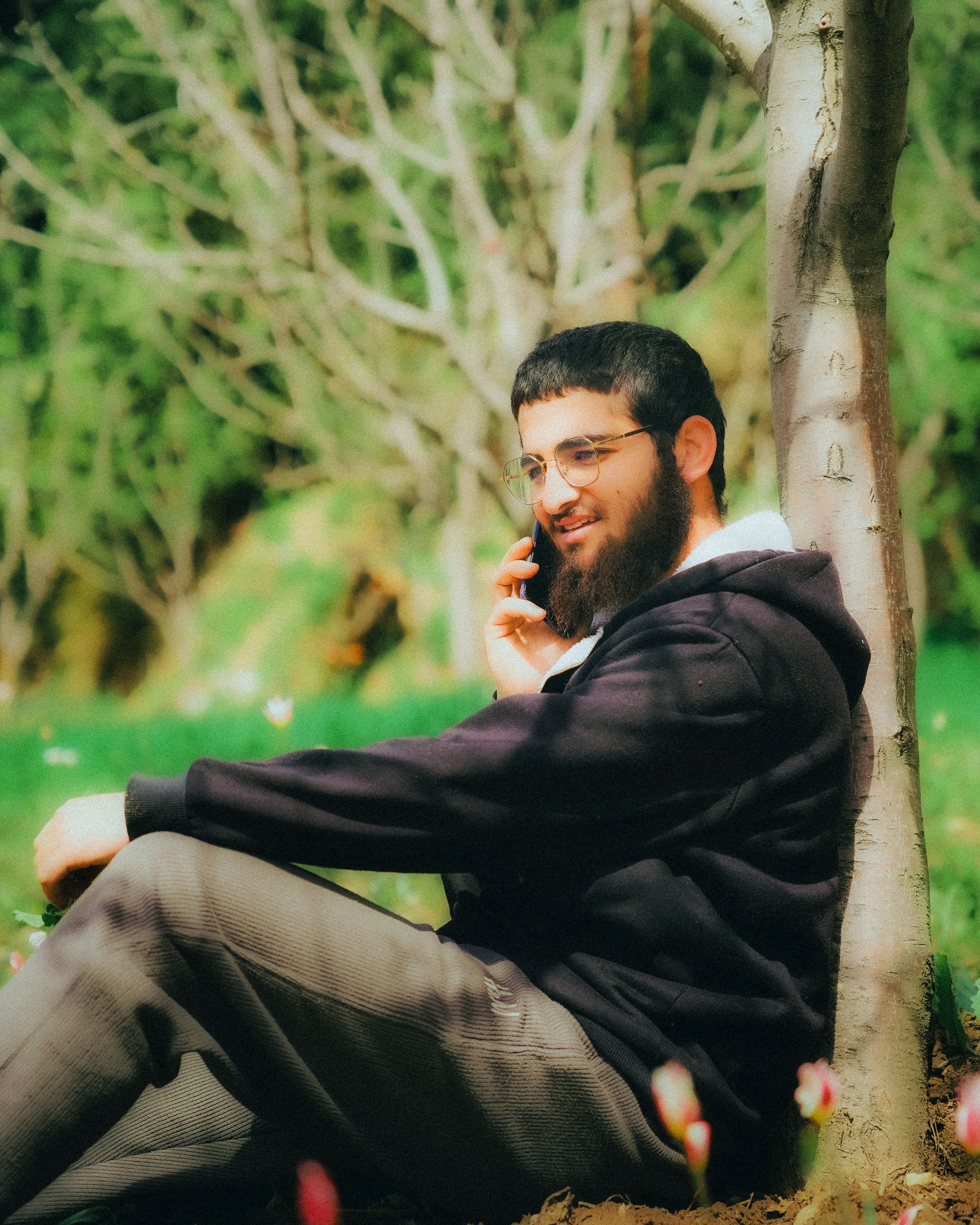 Adult man with beard and glasses sitting outdoors under a tree, speaking on his mobile phone.