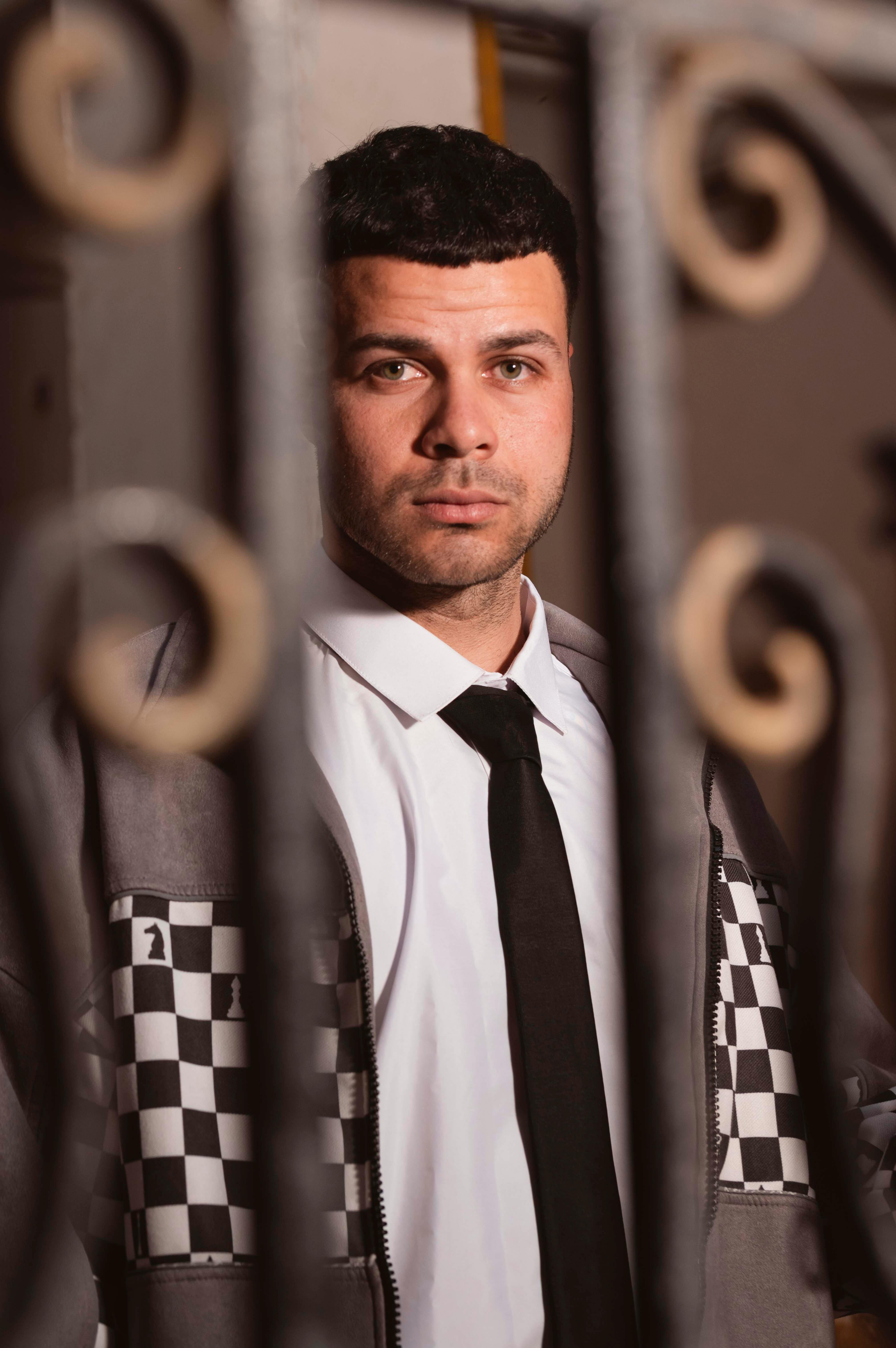 A man in a suit and checkered jacket stands confidently behind decorative metal bars.