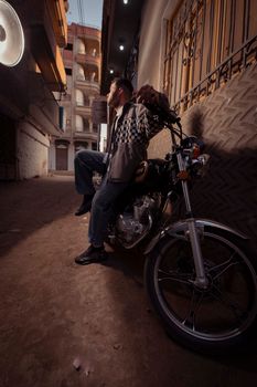 A man relaxes on his motorcycle in a narrow alley illuminated by streetlights.