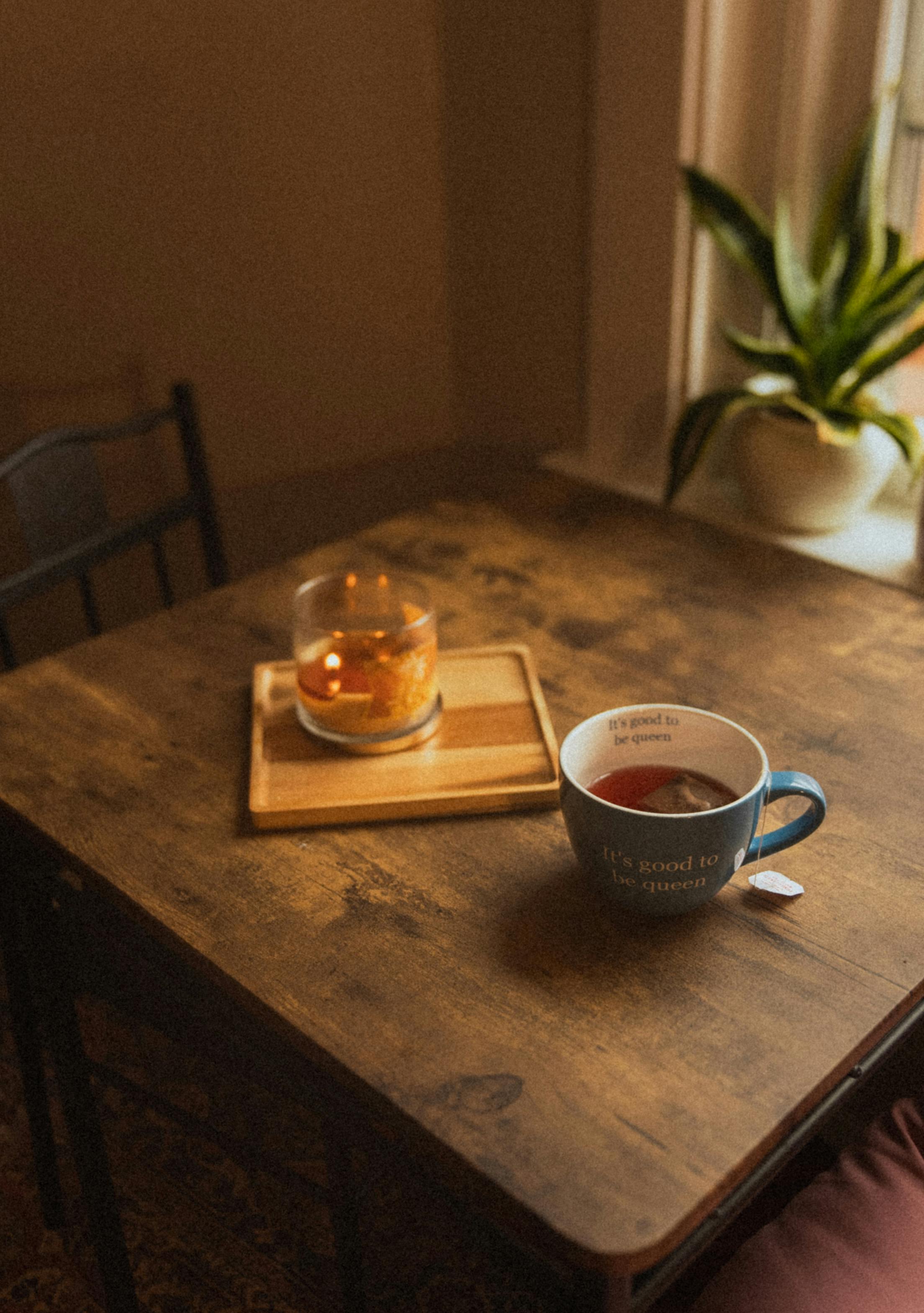 Warm and inviting scene of a tea cup and candle on a wooden table by a window with a plant.