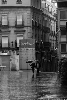 Black and white photo of people walking with umbrellas in a rainy Alicante alley.