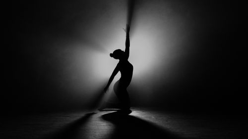 Free Silhouette of a dancer kneeling with arm raised, illuminated by a dramatic spotlight in a dark setting. Stock Photo