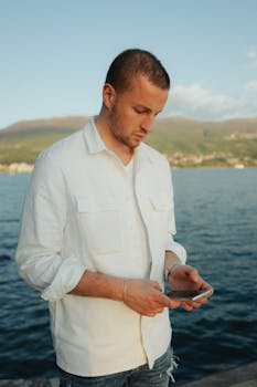 A young man in a white shirt using a smartphone near a tranquil sea backdrop.