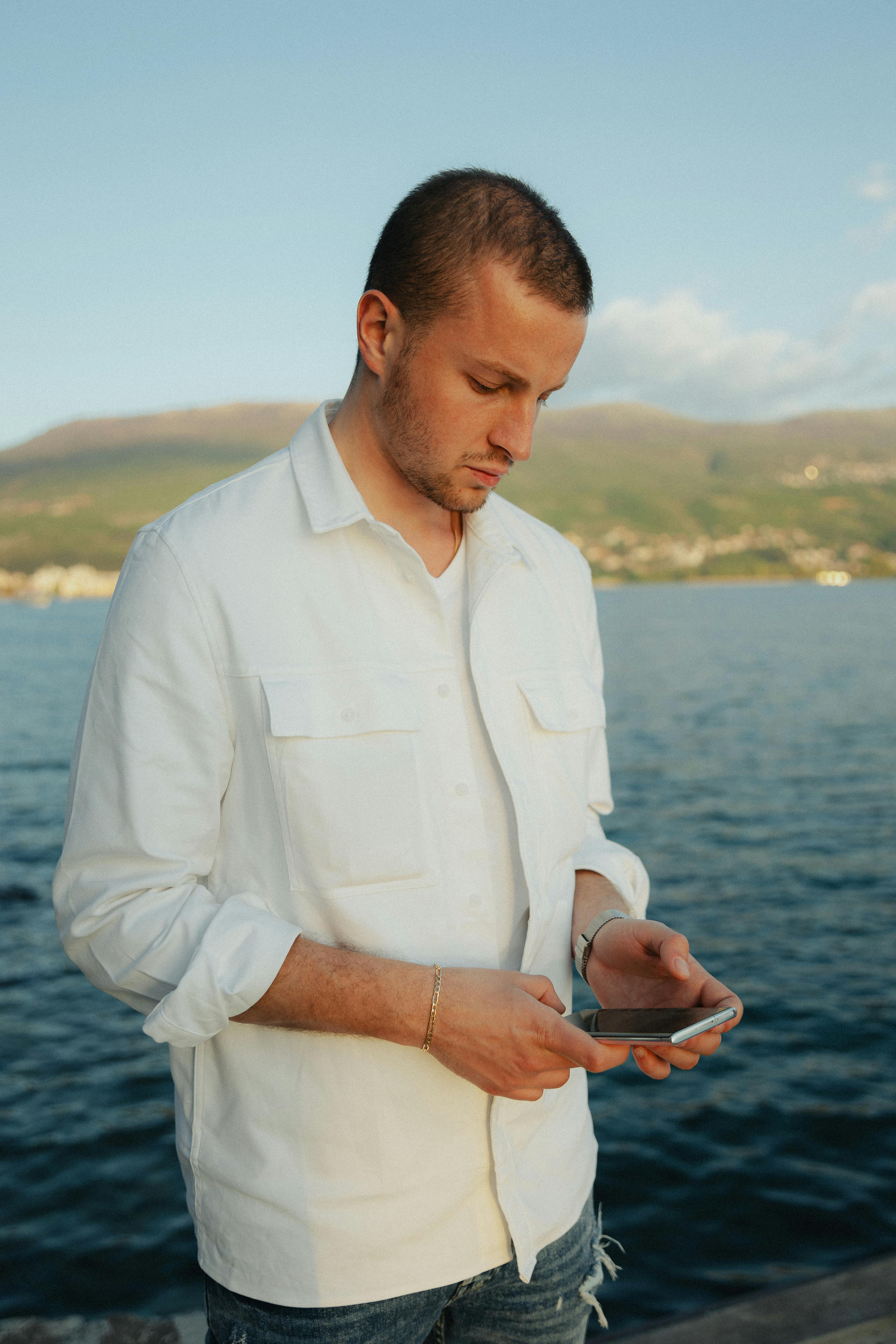 Free A young man in a white shirt using a smartphone near a tranquil sea backdrop. Stock Photo