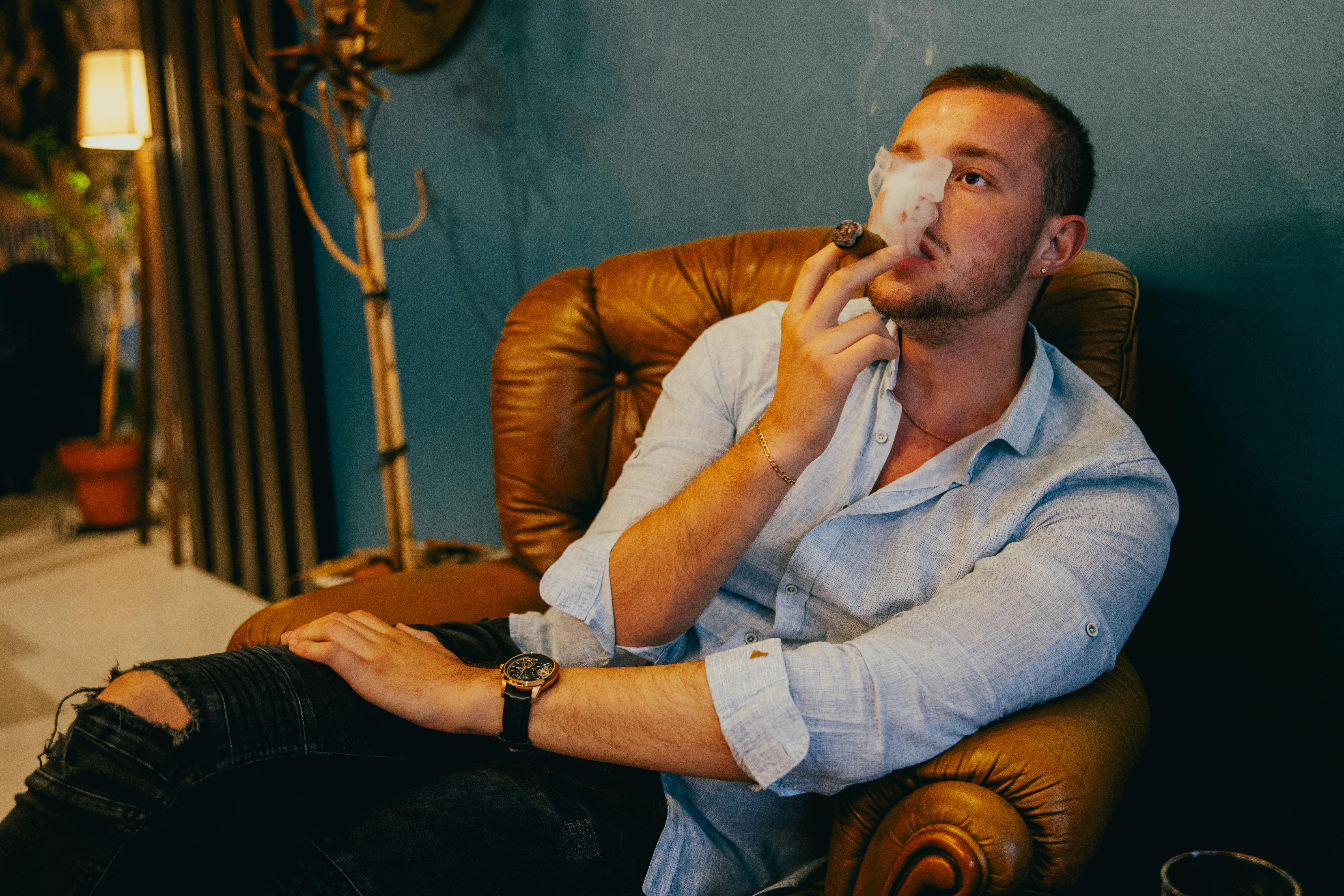 A man in a relaxed pose enjoying a cigar in a cozy armchair indoors.