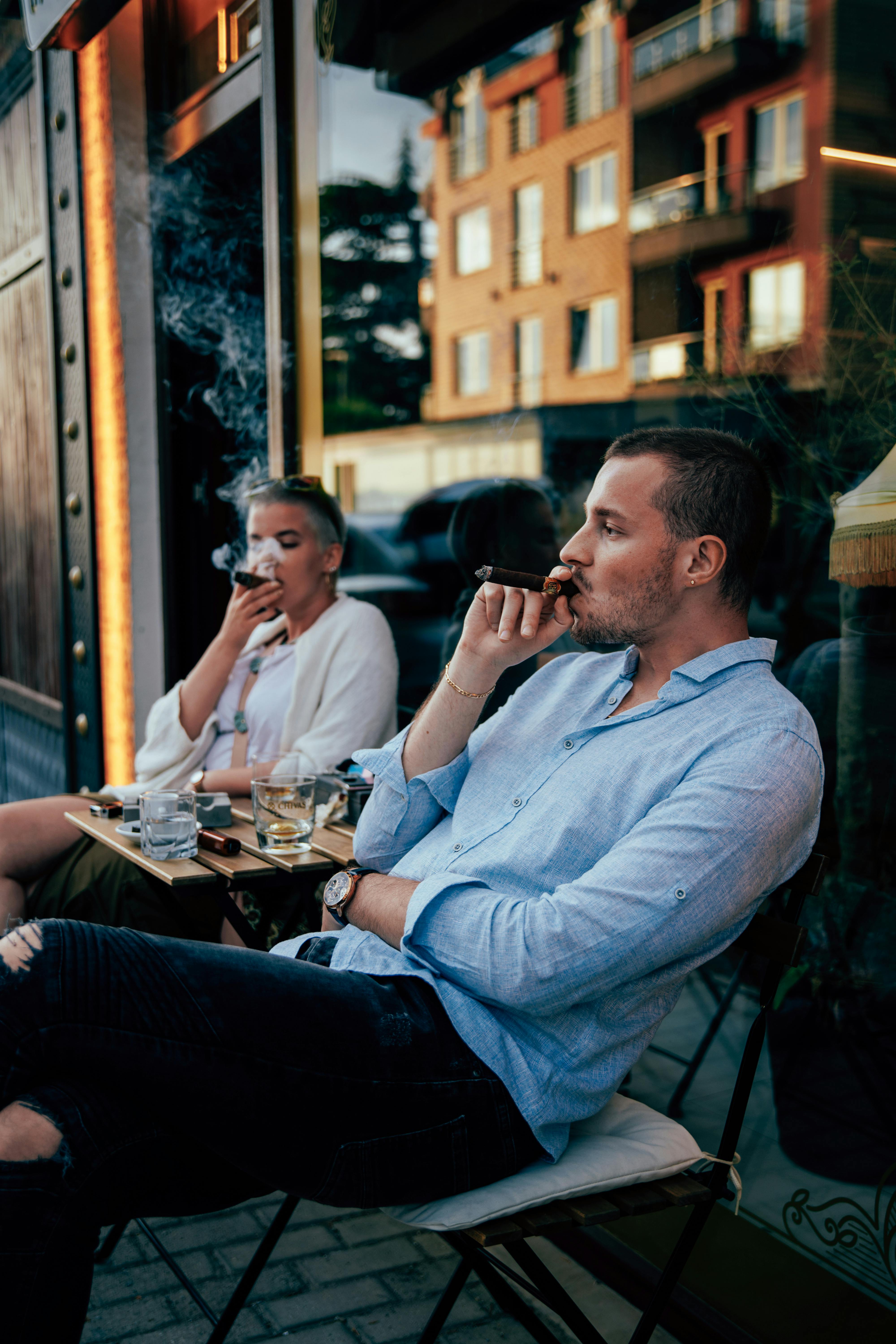 Two adults enjoying cigars and drinks outside a modern cafe on a summer evening.