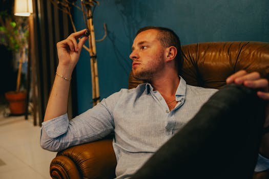 Man sitting in a leather armchair, holding a cigar in a relaxed indoor setting.