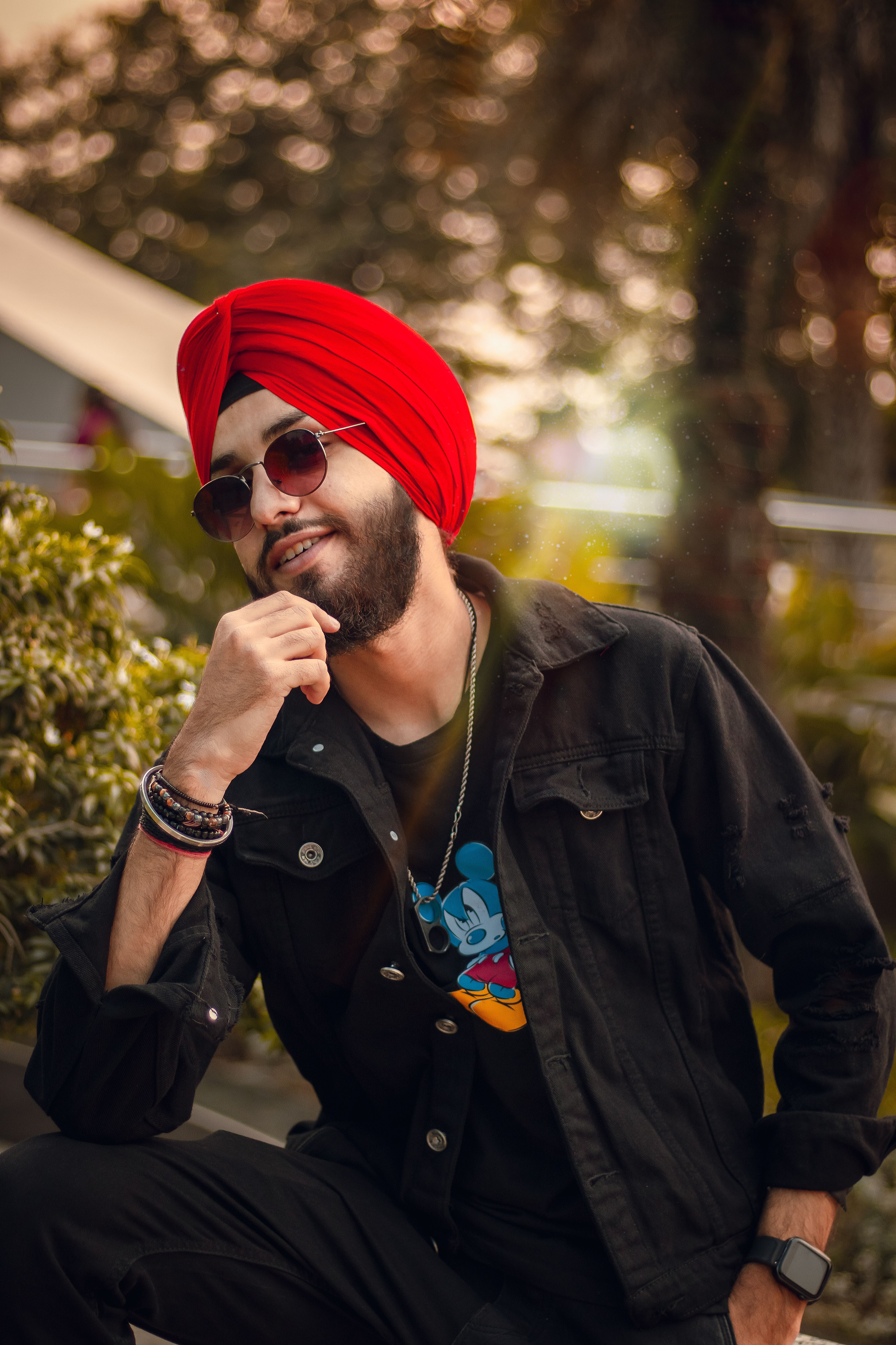 Smiling man in a red turban poses stylishly outdoors in a park setting.