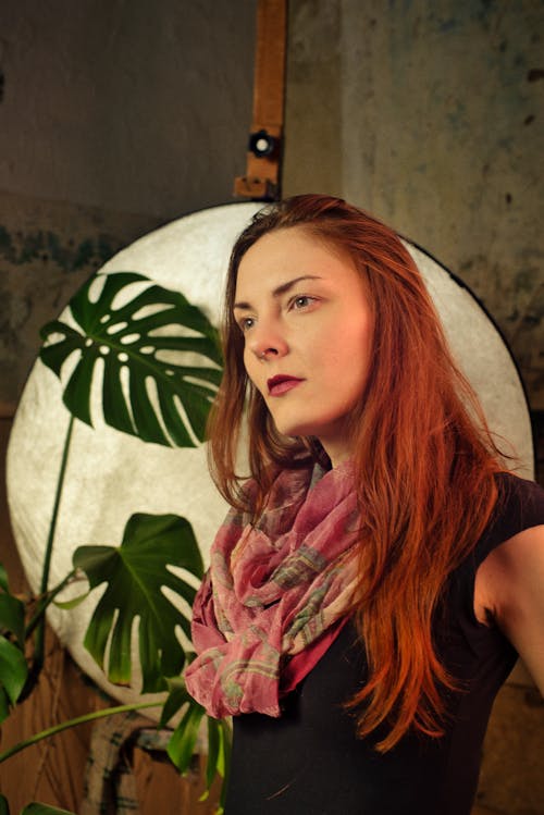 A woman with red hair and a scarf in front of a plant