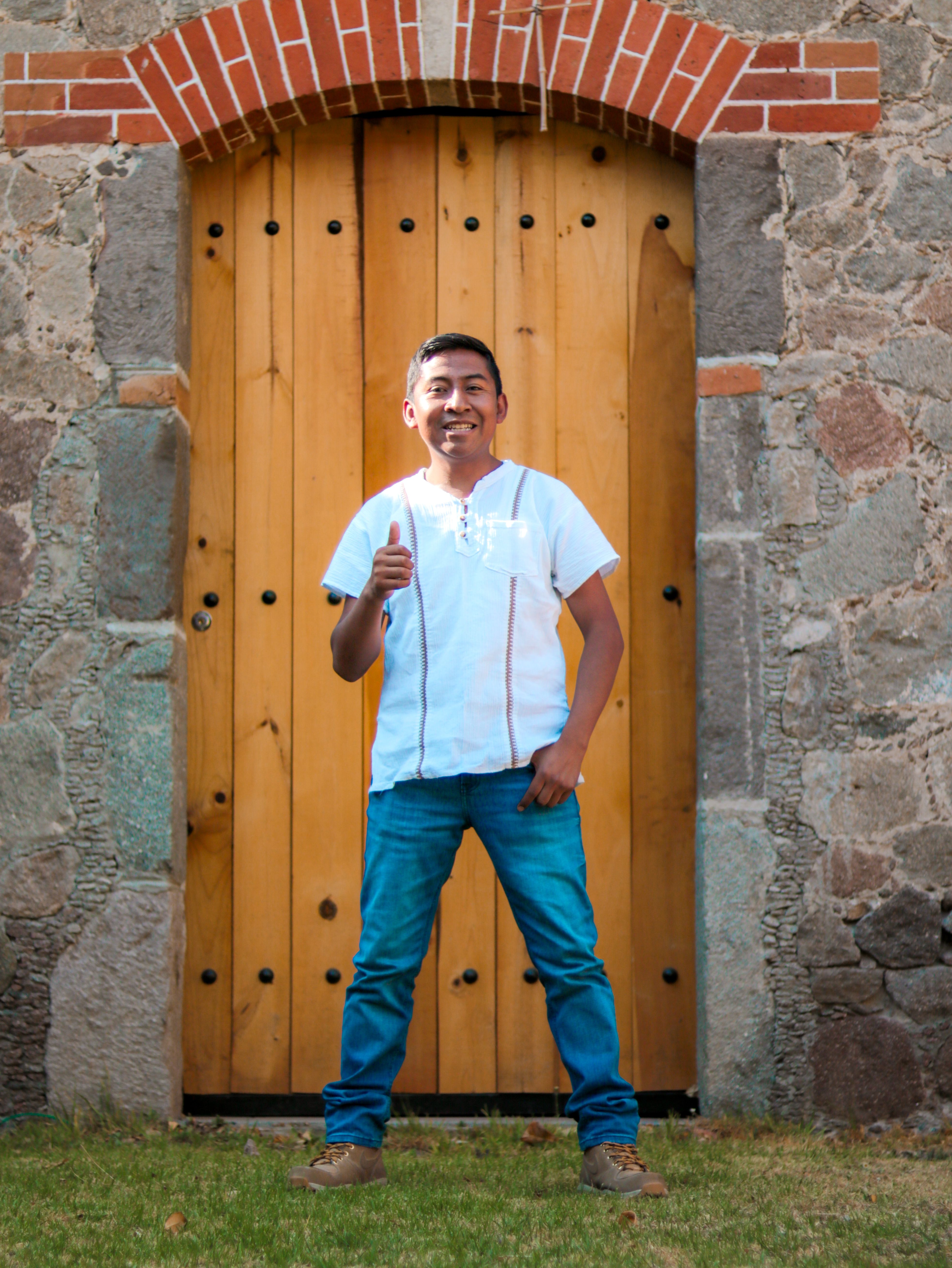 Confident man in casual attire posing with thumbs up in front of rustic wooden door.