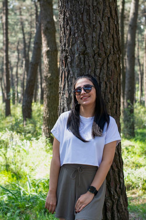 A woman in a white shirt and sunglasses standing next to a tree
