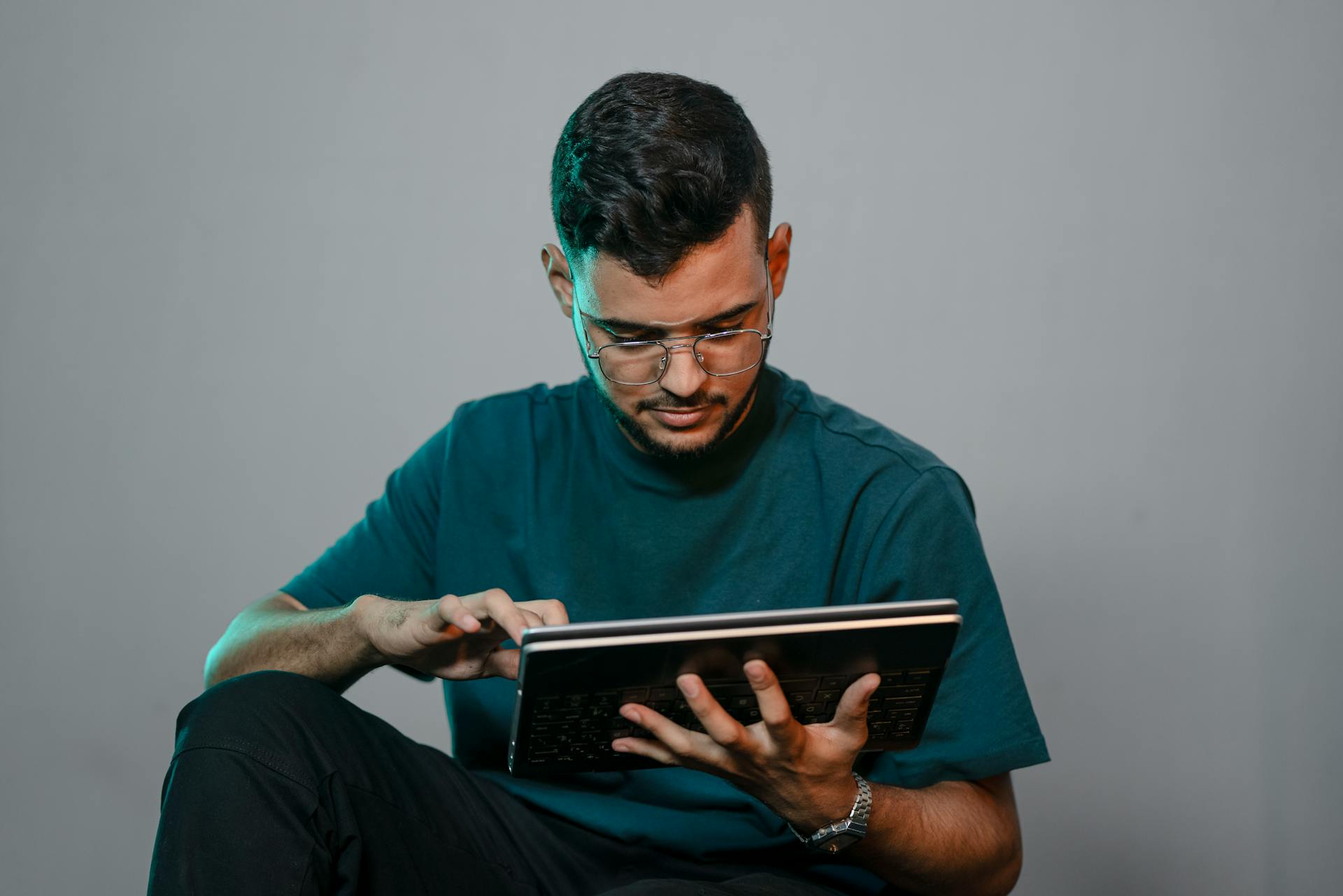 A young man with glasses intently scrolling on a tablet against a gray background.