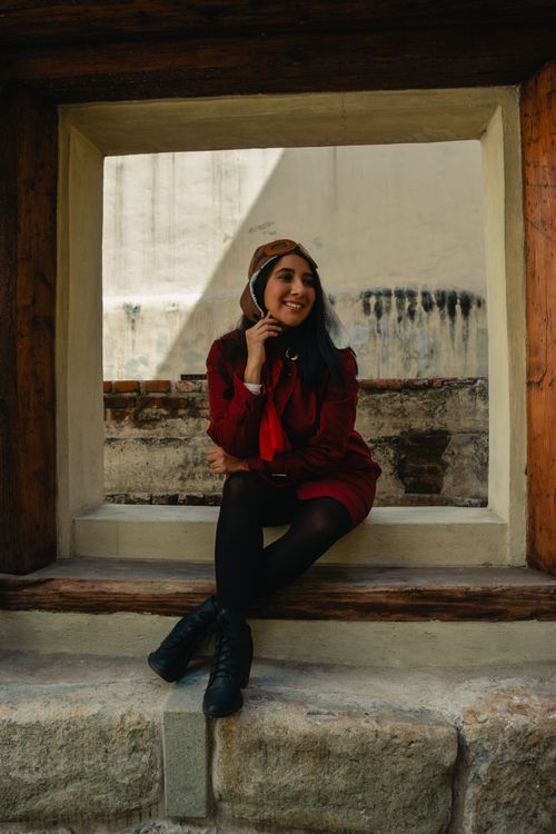 Free A stylish woman in a red jacket and aviator hat sits and smiles outside a historic building. Stock Photo