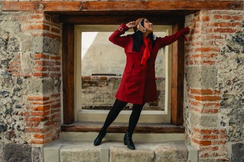 Free Young woman in a red coat poses playfully in Ciudad de México against an antique brick window frame. Stock Photo