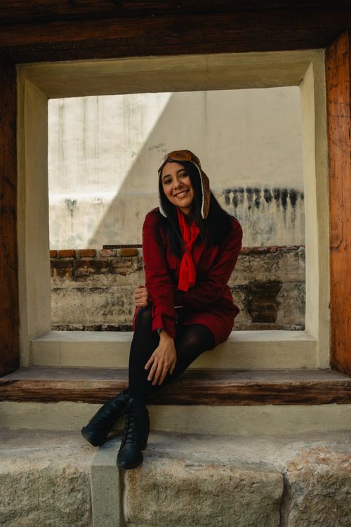 Free Cheerful woman in aviator hat and red coat posing by a window, exuding a stylish and happy vibe. Stock Photo