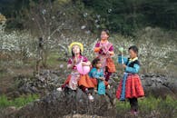 四个穿着色彩缤纷传统服装的孩子正在户外玩肥皂泡，周围风景优美