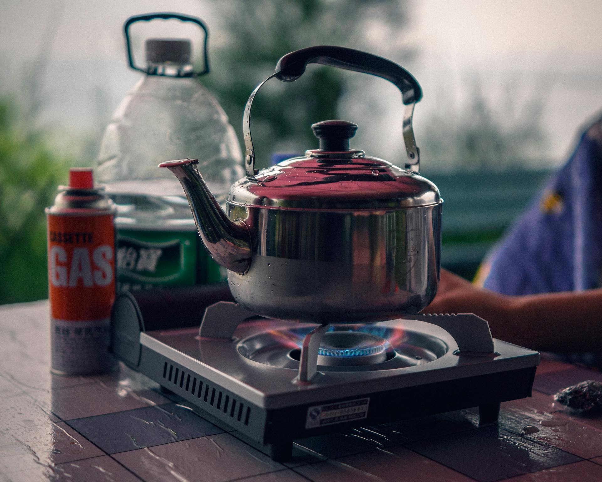 A kettle boils on an outdoor gas stove with visible steam, set on a wooden table.