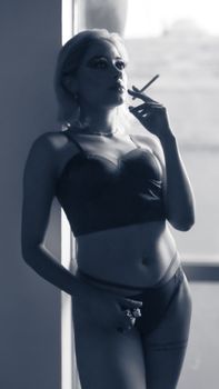 Sensual black and white portrait of a young woman smoking indoors in lingerie.