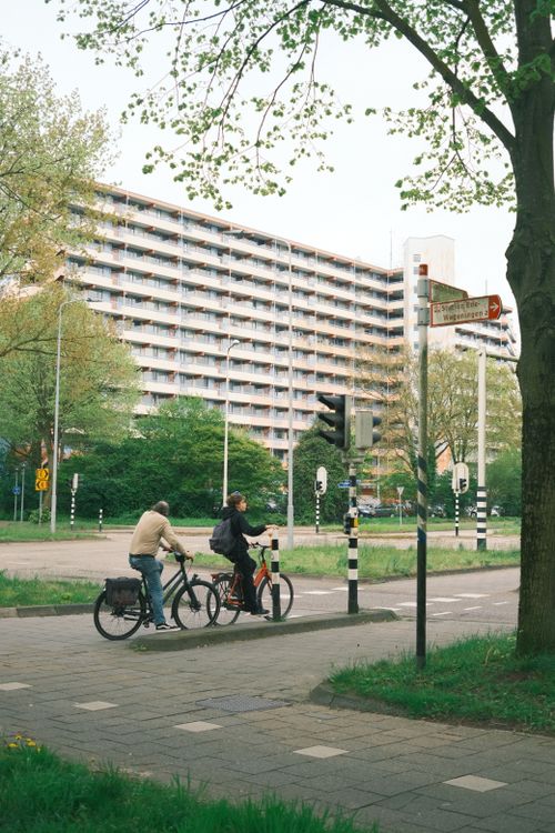 Free Two cyclists pause at a street crossing in Amsterdam against a backdrop of urban architecture. Stock Photo