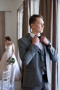 A groom adjusts his tie in a refined suit as a bride waits in the background, creating a classic wedding scene.