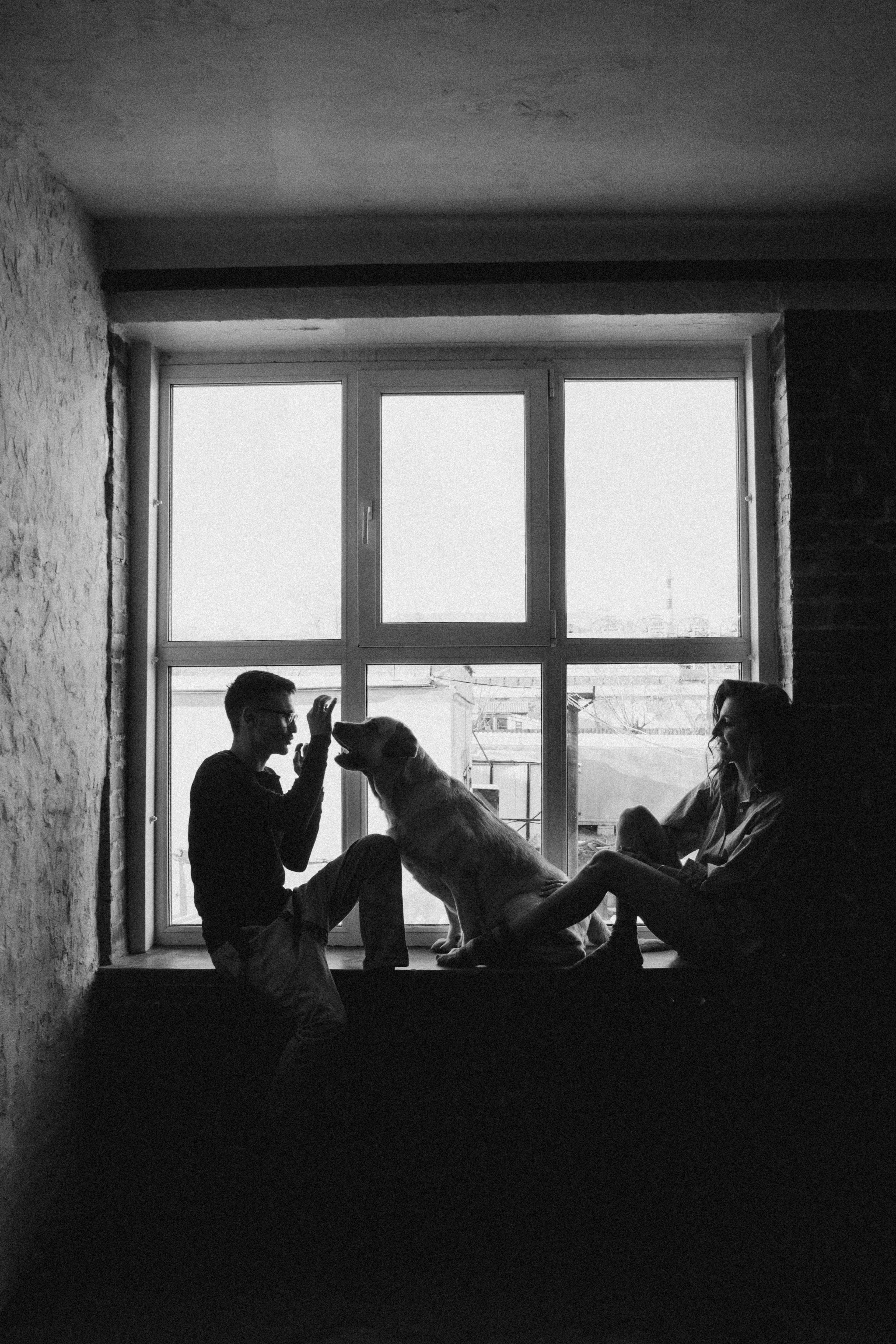 Monochrome portrait of a couple and their dog sitting by a window in an urban setting.