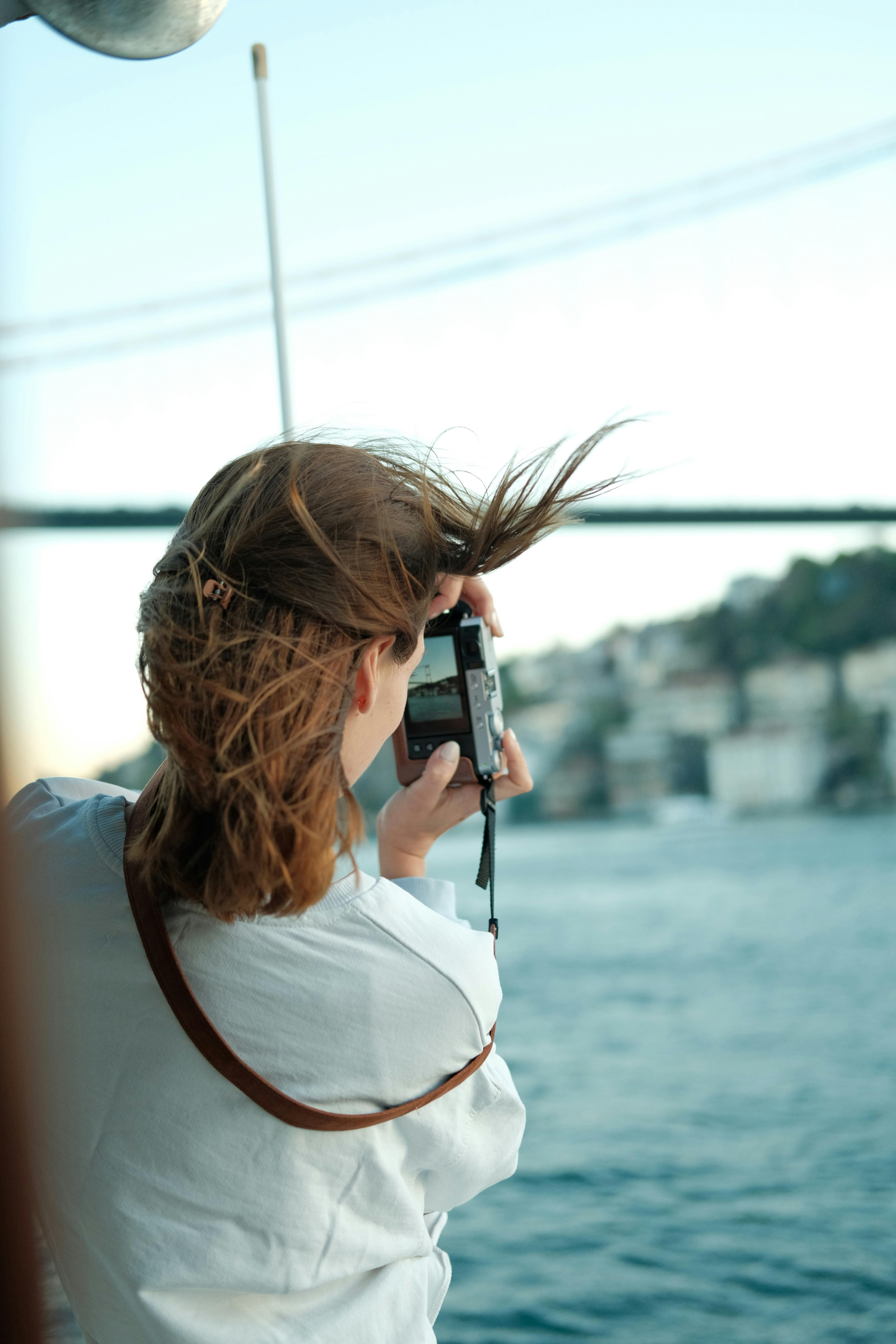 A woman takes a photo by the sea with her hair blowing in the wind, capturing a scenic view.