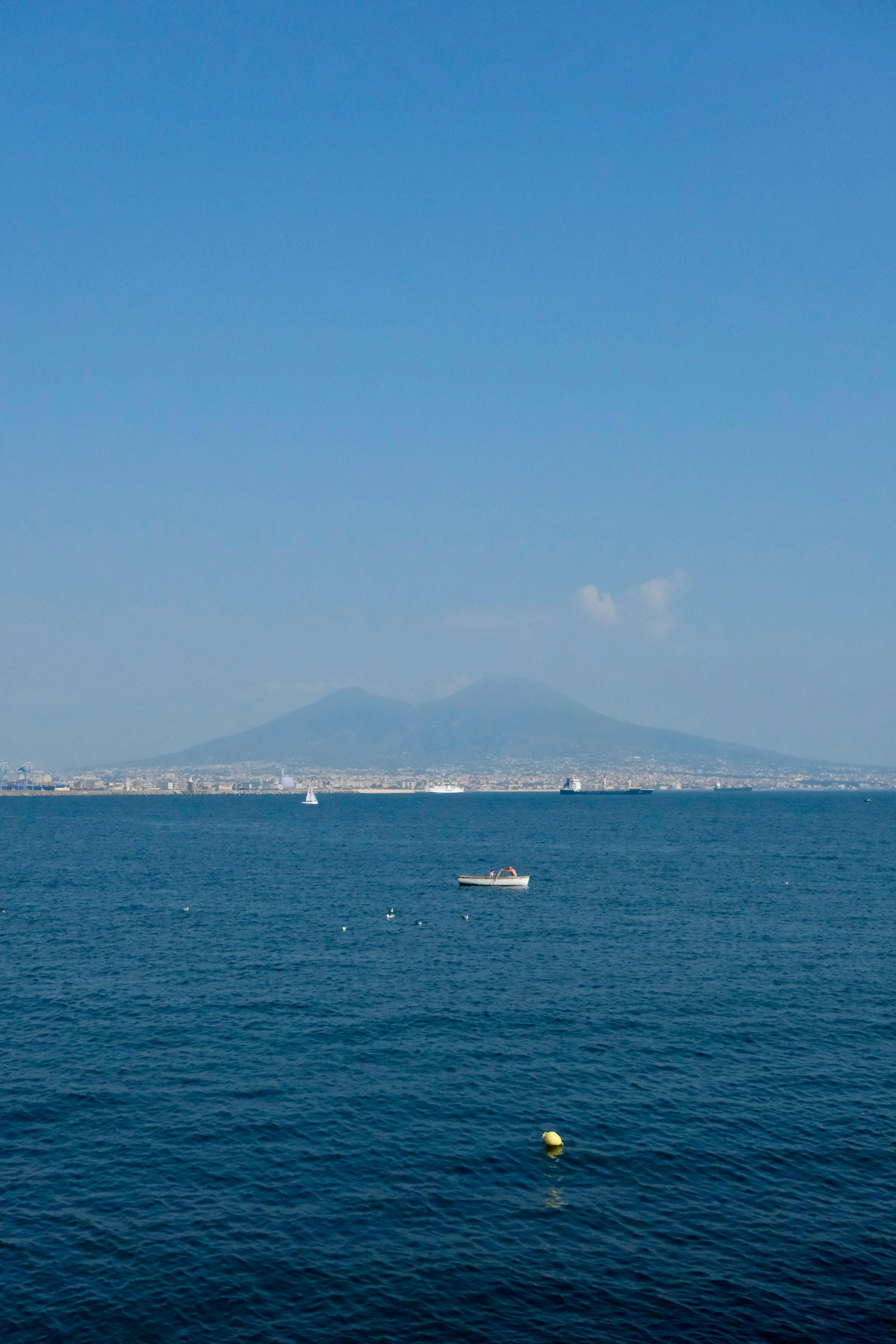 A serene view of Mount Vesuvius across the bay from Napoli on a clear day, featuring small boats.