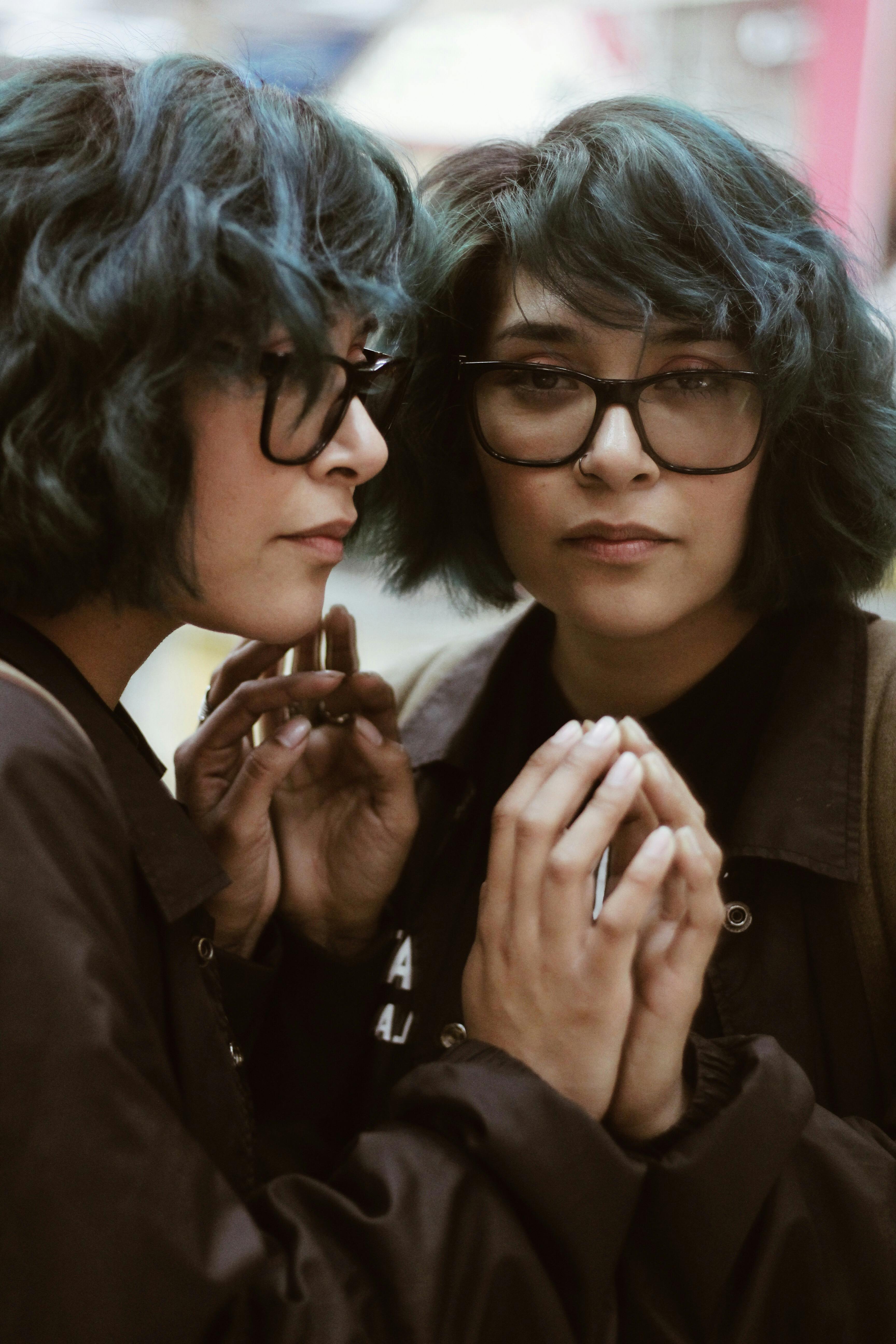 留着青色头发、戴着眼镜的时尚女人凝视着镜子，倒影中透出深思的神情