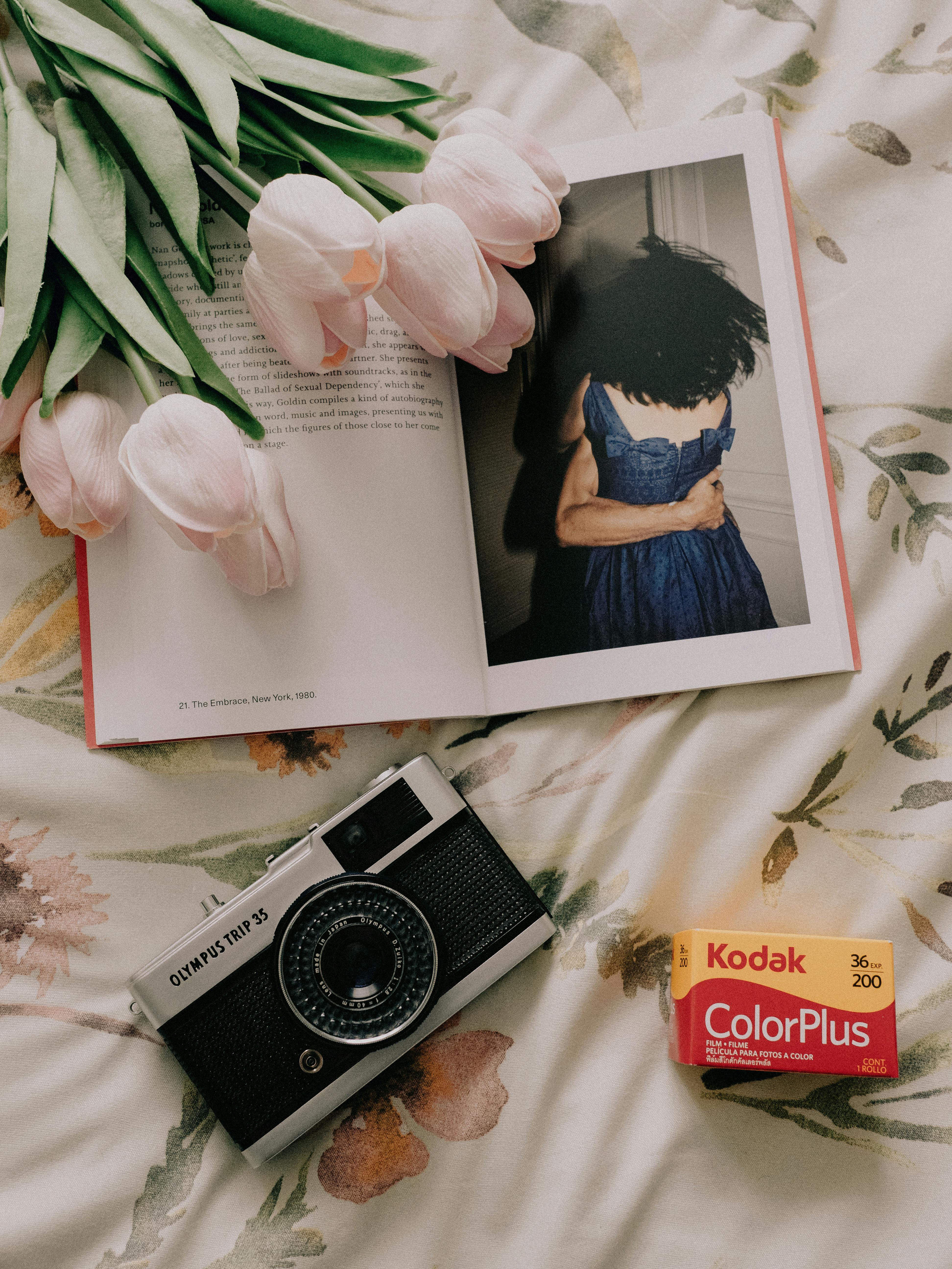 Top view of a vintage camera, tulips, and an open book on a floral background, accompanied by a film roll box.