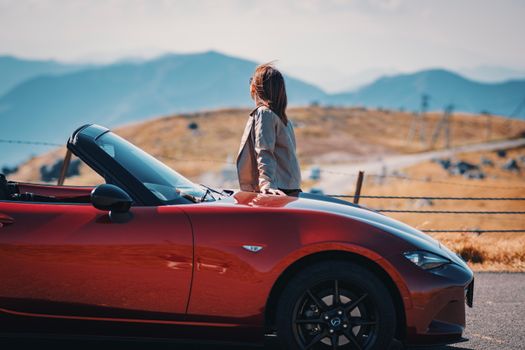 A woman standing by a red convertible sports car, admiring a scenic mountain view.