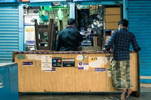 Two men at a shop counter in Bengaluru marketplace with payment signs and products displayed.