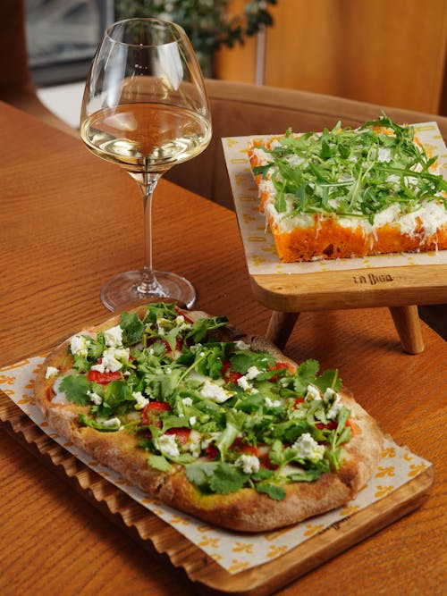 A Glass of White Wine Standing on a Table next to a Pinsa and a Dish with Arugula 