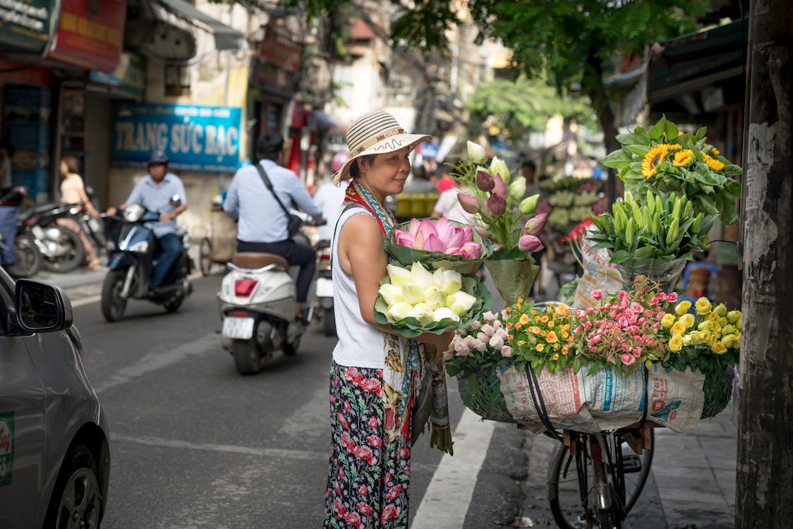 Free A woman sells vibrant flower bouquets on a bicycle in a bustling city street market. Stock Photo