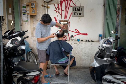 Street barber gives haircut in urban setting with motorbikes nearby.
