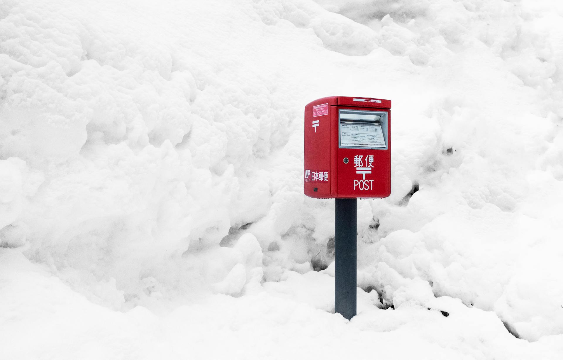 A red Japanese mailbox surrounded by thick winter snow, creating a striking contrast.