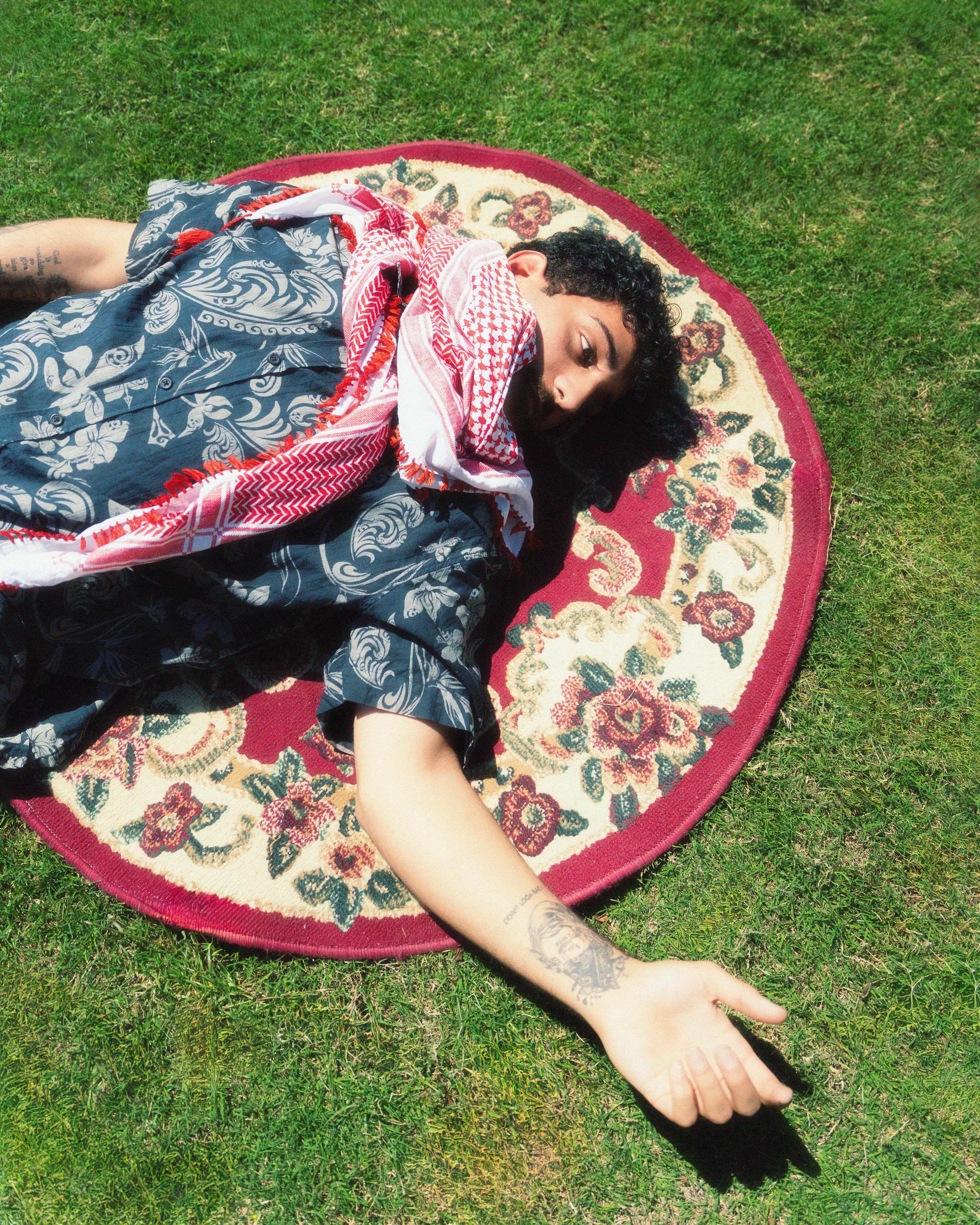 A stylish man with tattoos lies on a floral carpet over grass, showcasing casual fashion and unique style.