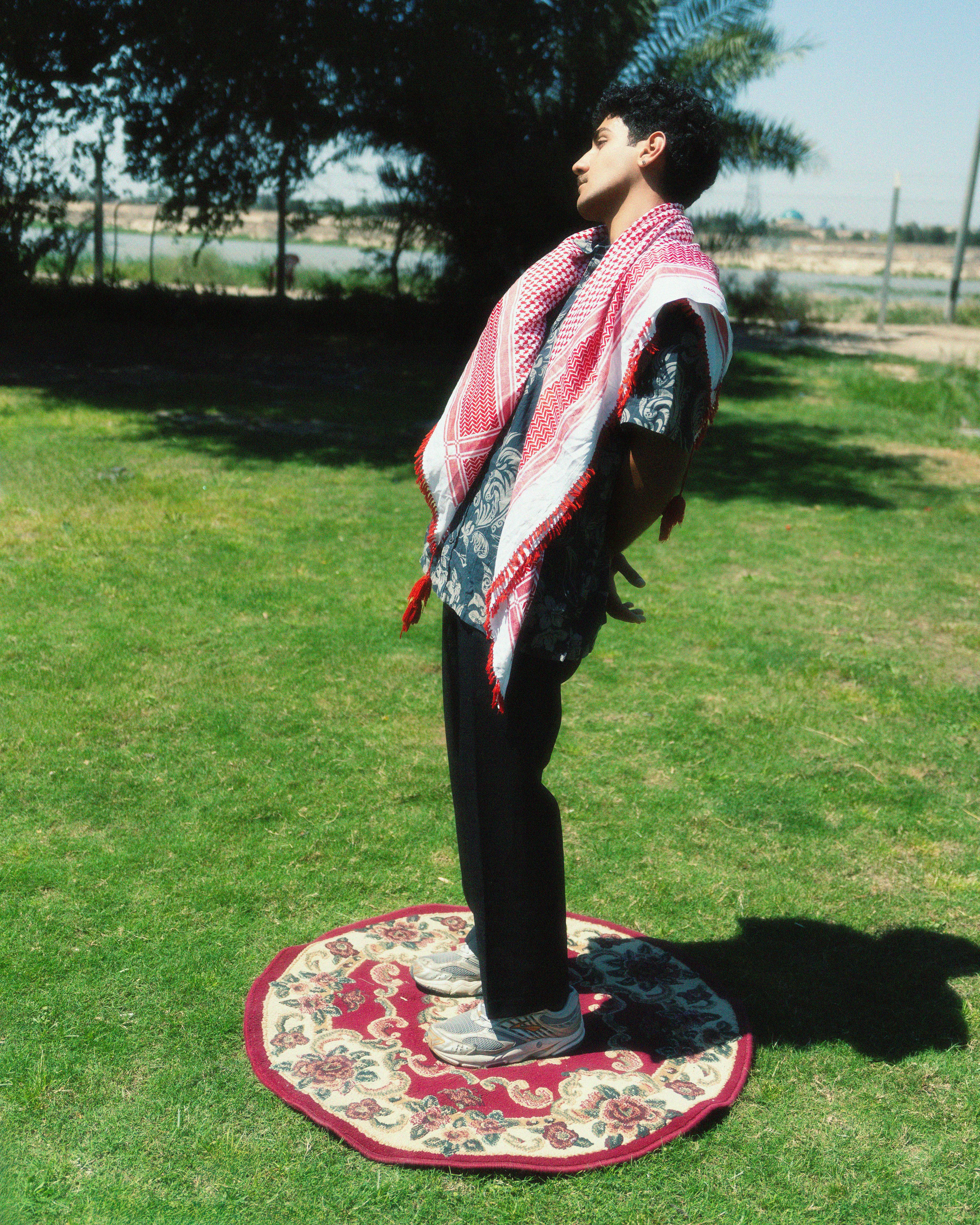 Man wearing traditional shawl standing on carpet outdoors in a sunny, rural area.