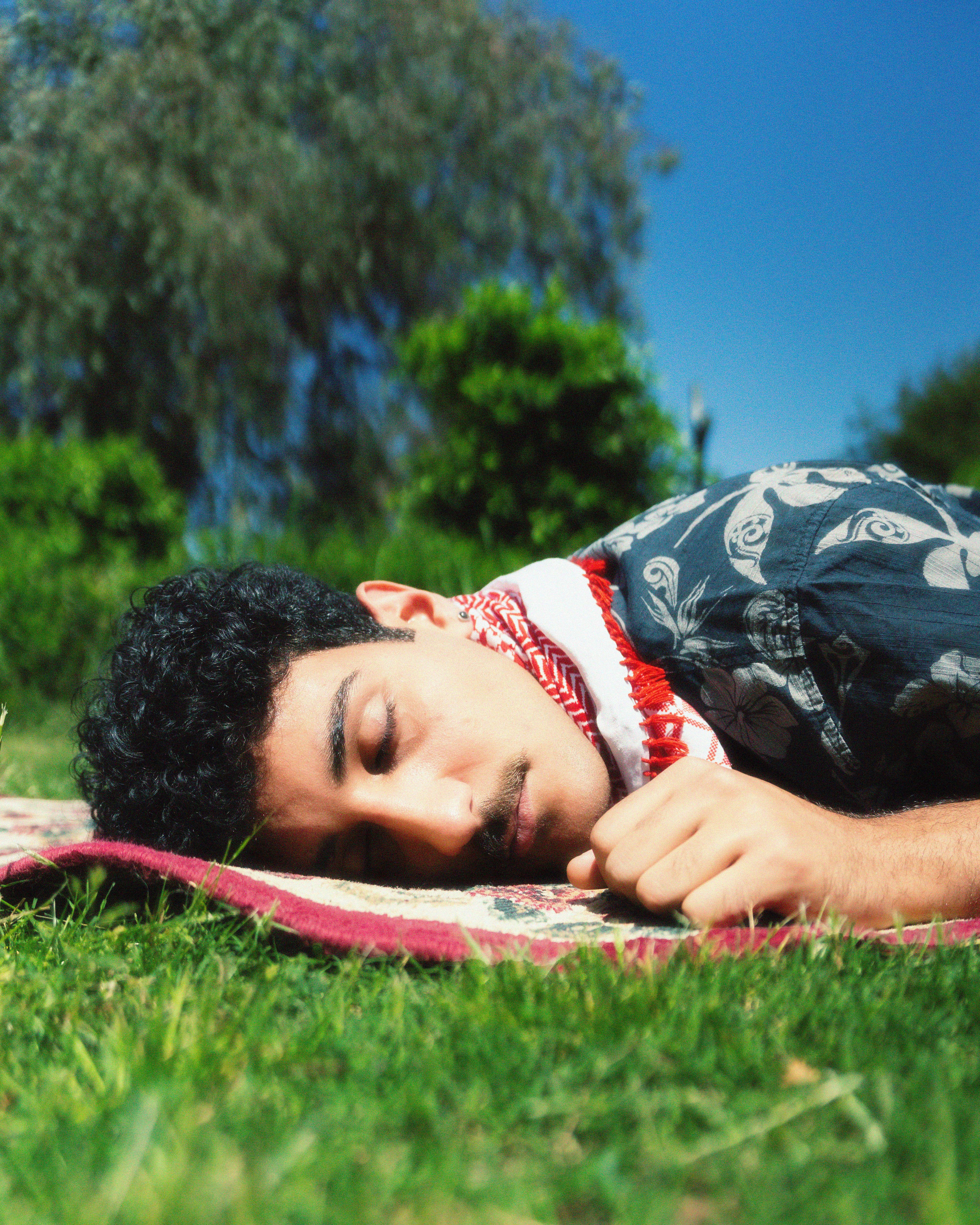 A man with short hair lies peacefully on a carpet on grass under a clear blue sky.